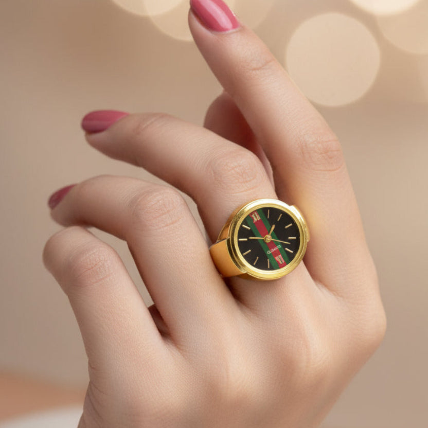 A gold-tone ring designed to look like a Gucci watch, featuring a black dial with green and red stripes, held in a small pink round presentation box.
